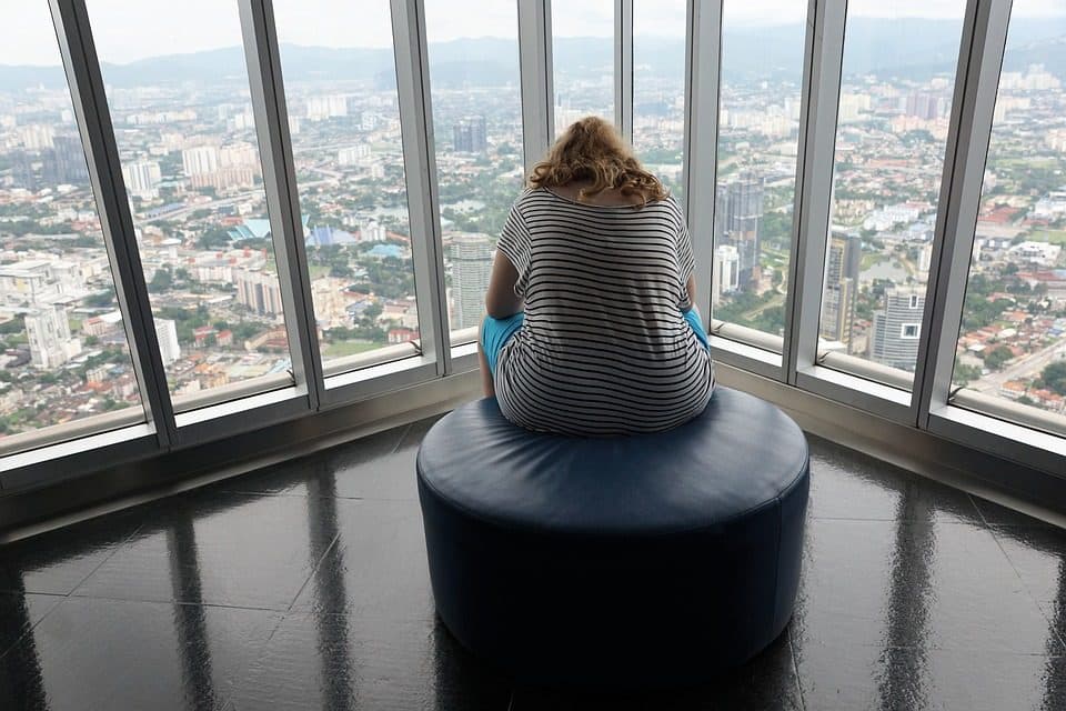 Femeie așezată cu spatele, privind orașul de la fereastra unei clădiri înalte