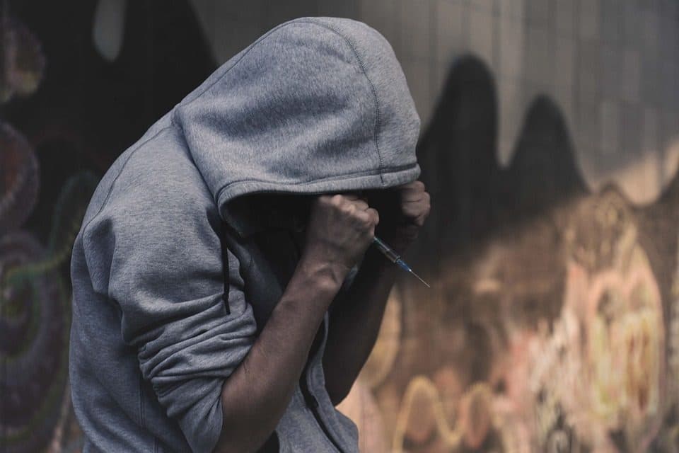 Persoană cu glugă ținând o seringă, lângă un perete cu graffiti