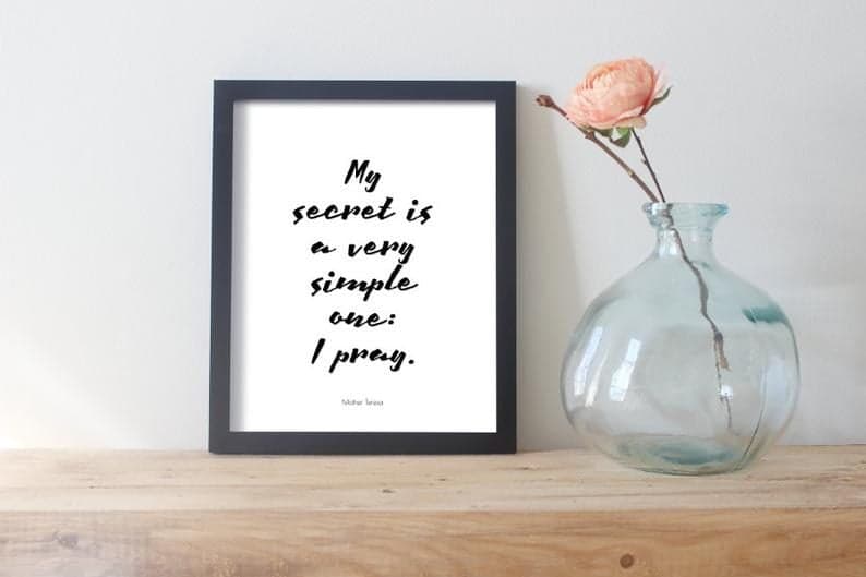 Tablou înrămat cu textul „My secret is a very simple one: I pray." lângă o vază cu un trandafir
