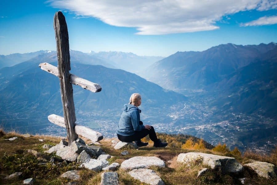 Persoană așezată lângă o cruce de lemn, privind spre o vale montană