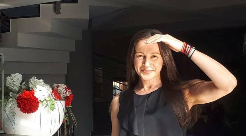 Femeie zâmbind, cu mâna la frunte, lângă un ghiveci cu flori albe și roșii