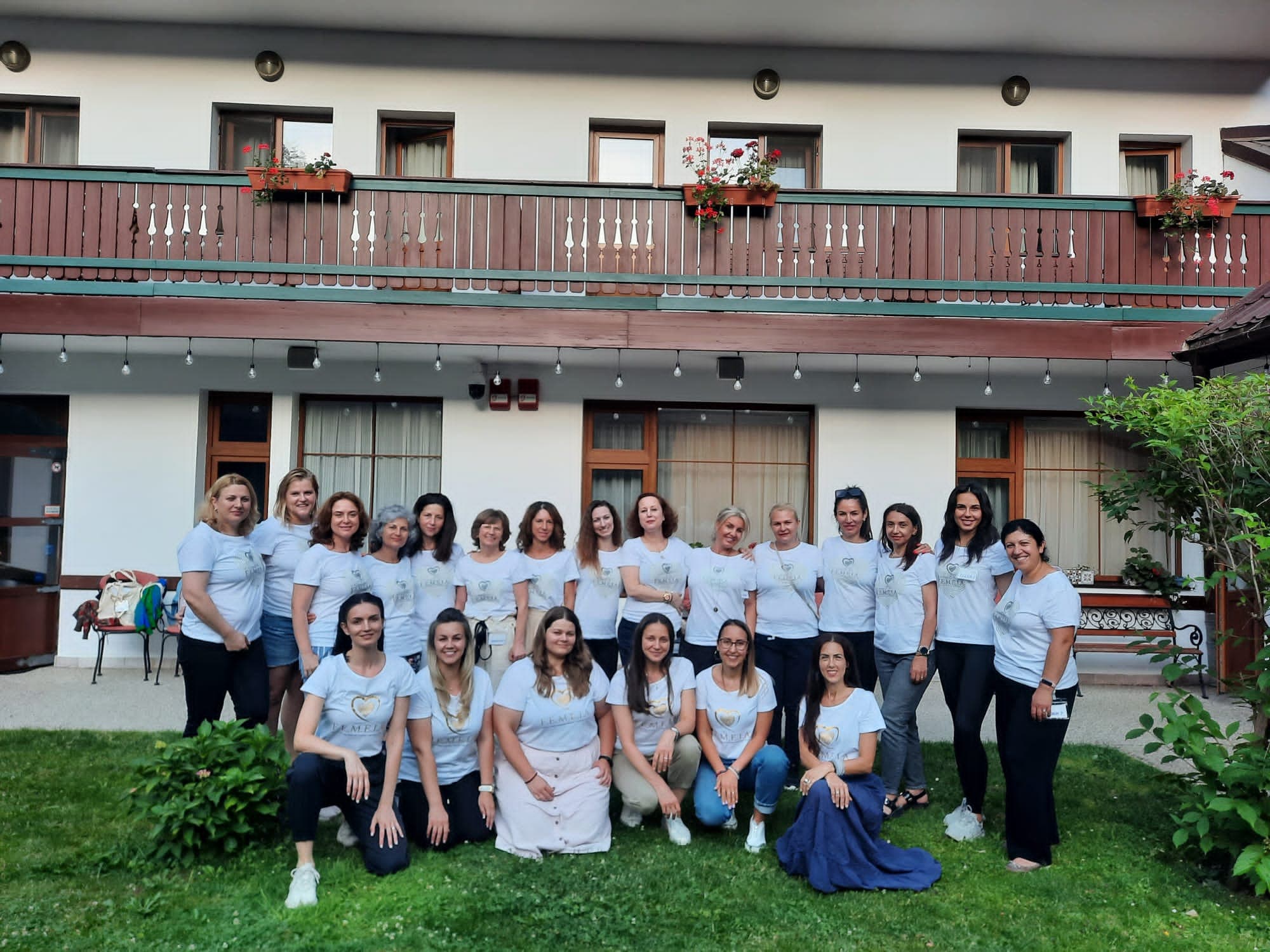 Grup de femei în tricouri albe, în fața unei clădiri cu balcon de lemn