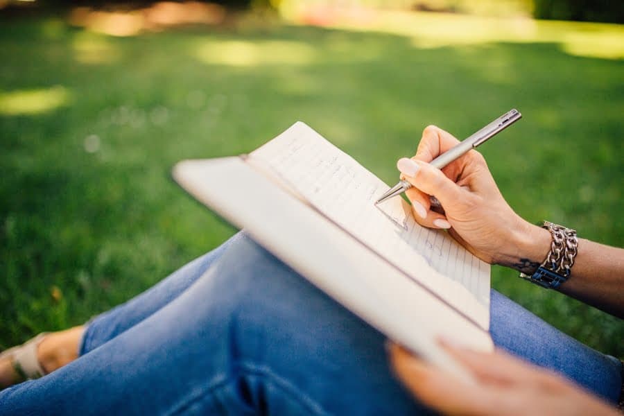 Mână de femeie scriind într-un caiet, așezată pe iarbă