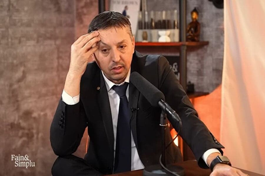 Bărbat în costum, cu mâna la frunte, la microfonul emisiunii „Fain&Simplu”