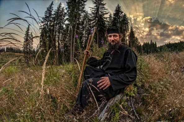 Bărbat în haină monahală neagră, cu toiag, într-o poiană de munte la apus