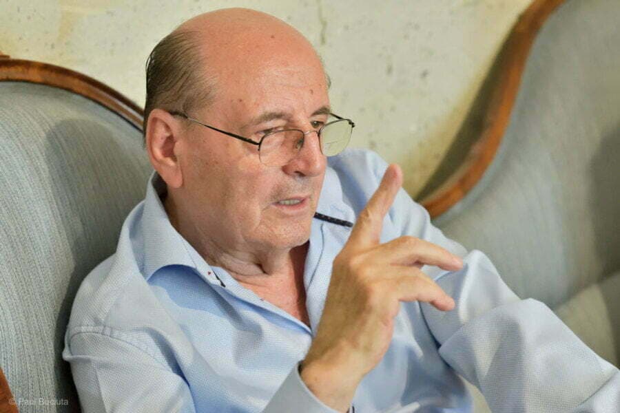 Bărbat în vârstă cu ochelari, într-un fotoliu, gesticulând cu un pix