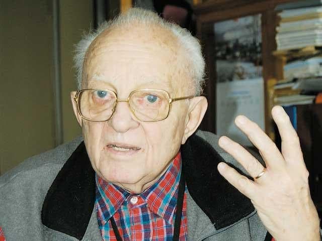 Bărbat vârstnic cu ochelari vorbind și gesticulând cu mâna