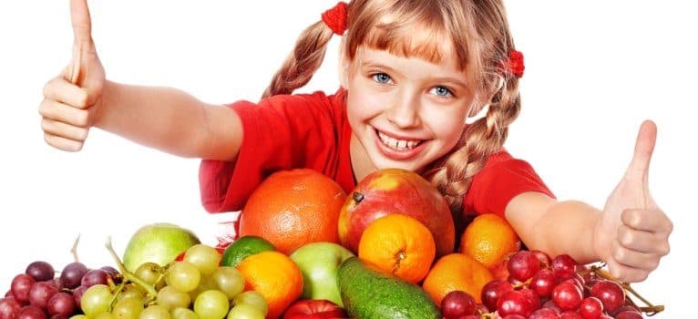 Fetiță zâmbind cu degetele mari ridicate, în spatele unui morman de fructe