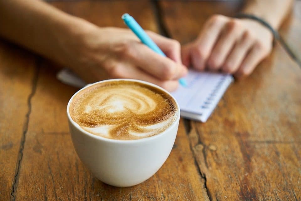 Ceașcă de cafea cu inimă în spumă, lângă cineva care scrie