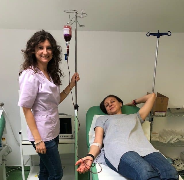 Femeie în uniformă roz zâmbind lângă o pacientă cu perfuzie