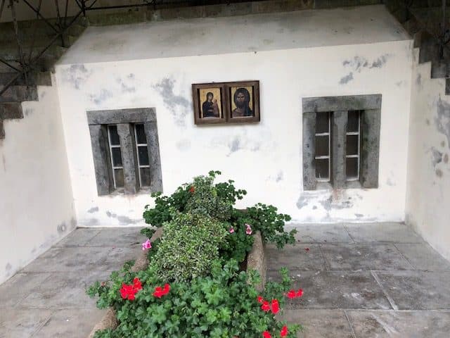 Curte cu două icoane pe un perete alb și mușcate înflorite