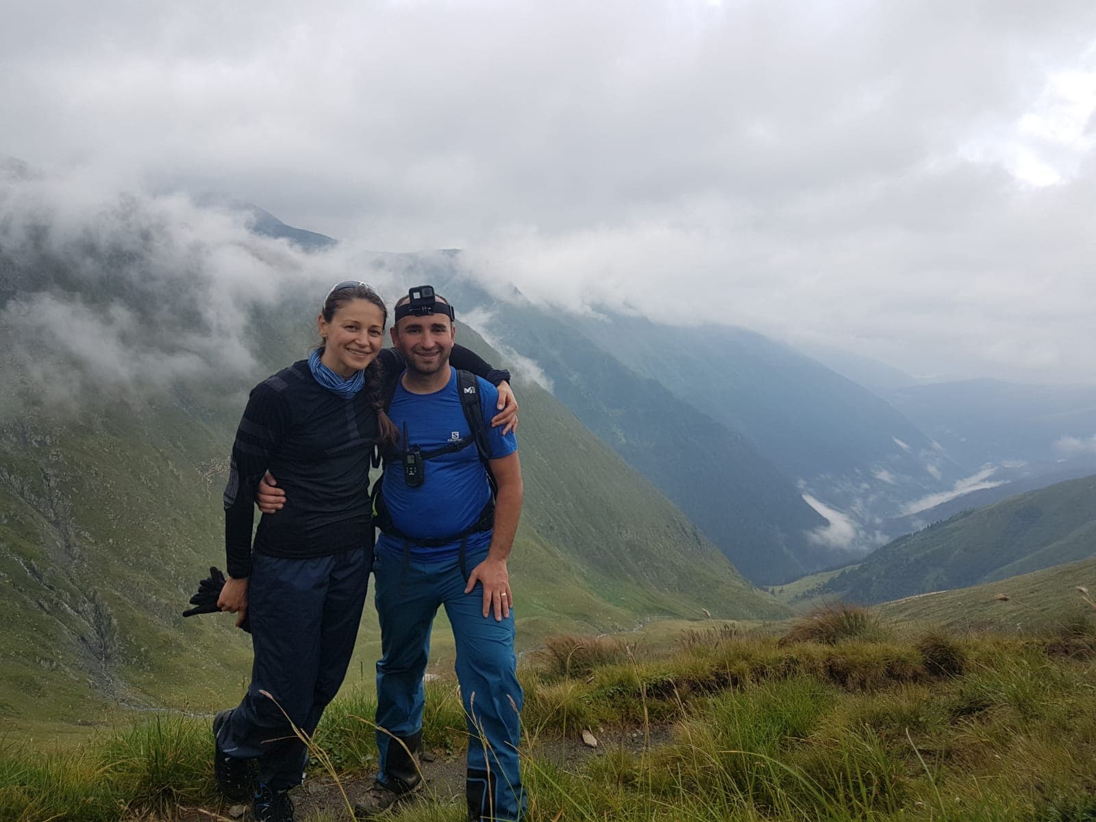 Femeie și bărbat îmbrățișați pe o culme de munte, cu nori în vale