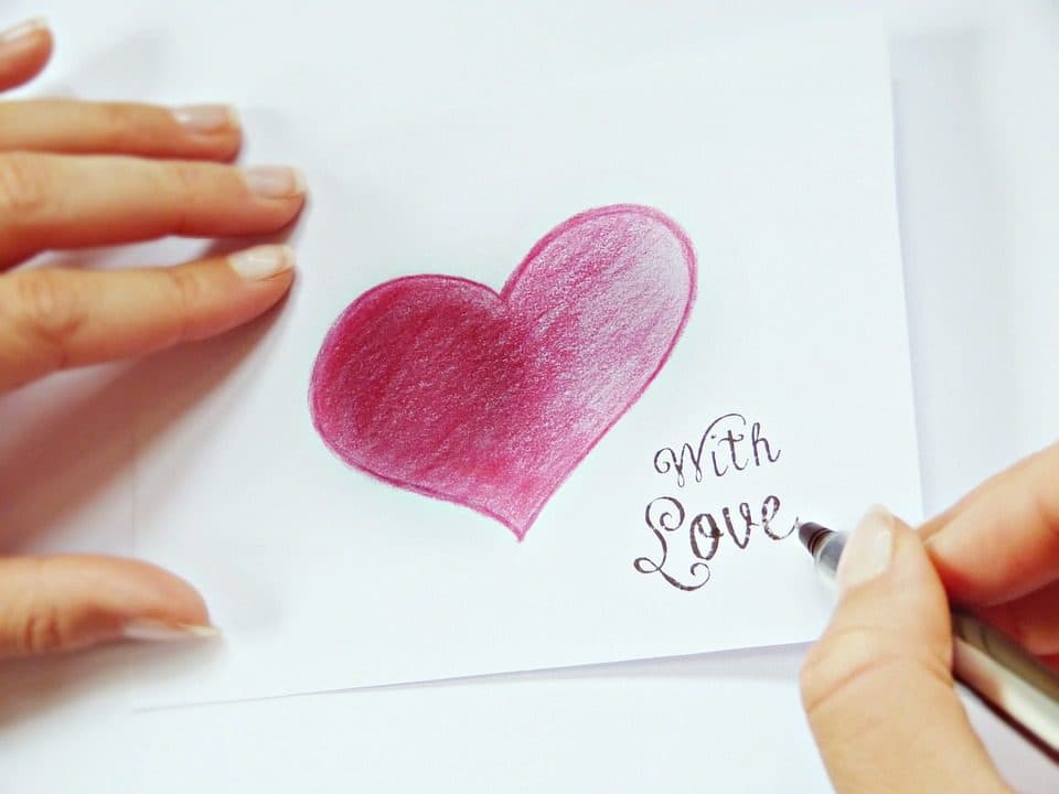 Mâini desenând o inimă roz lângă textul scris de mână „With Love”