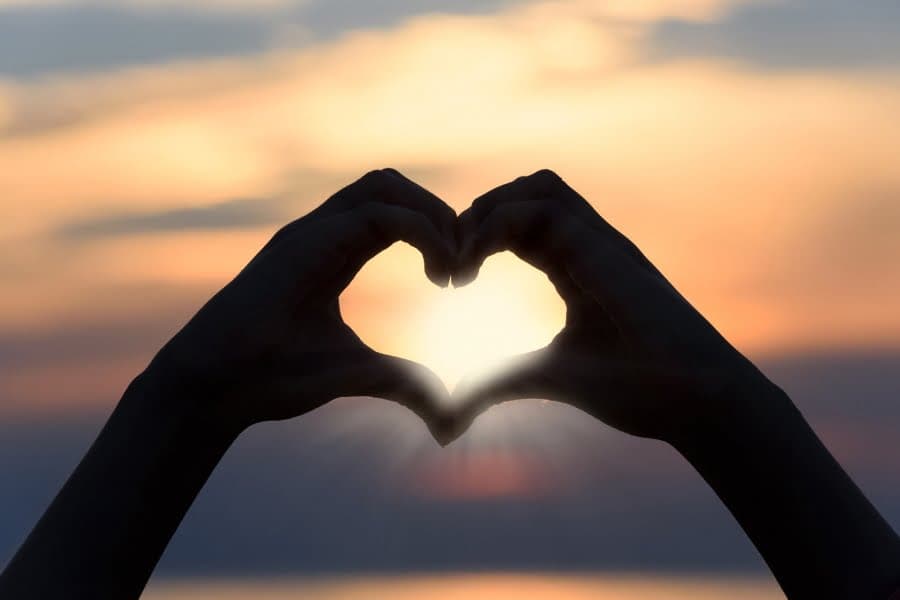 Mâini care formează o inimă în silueta apusului deasupra mării